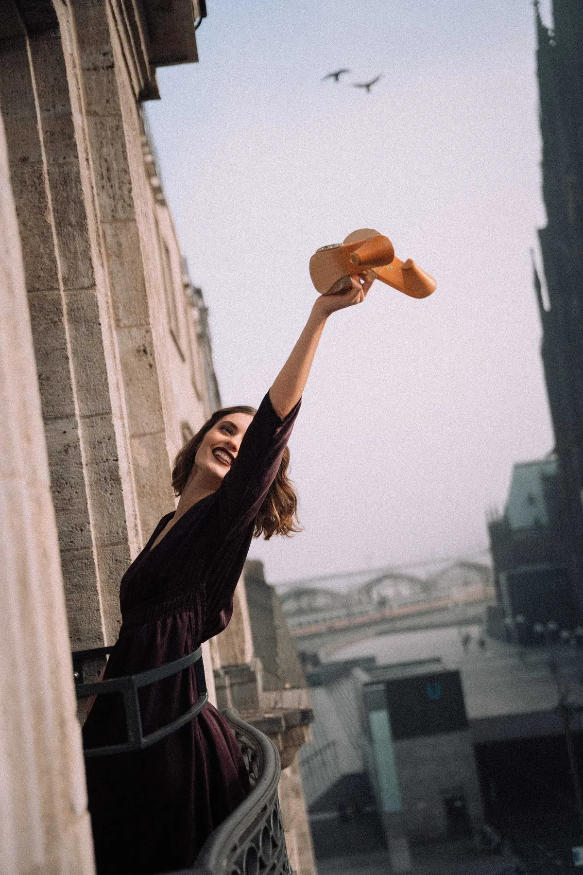 The Wants, model in a dark dress leaning out of a window holding a clog up to the sky, birds overhead, campaign 2020, photographed by Kilian Amrehn