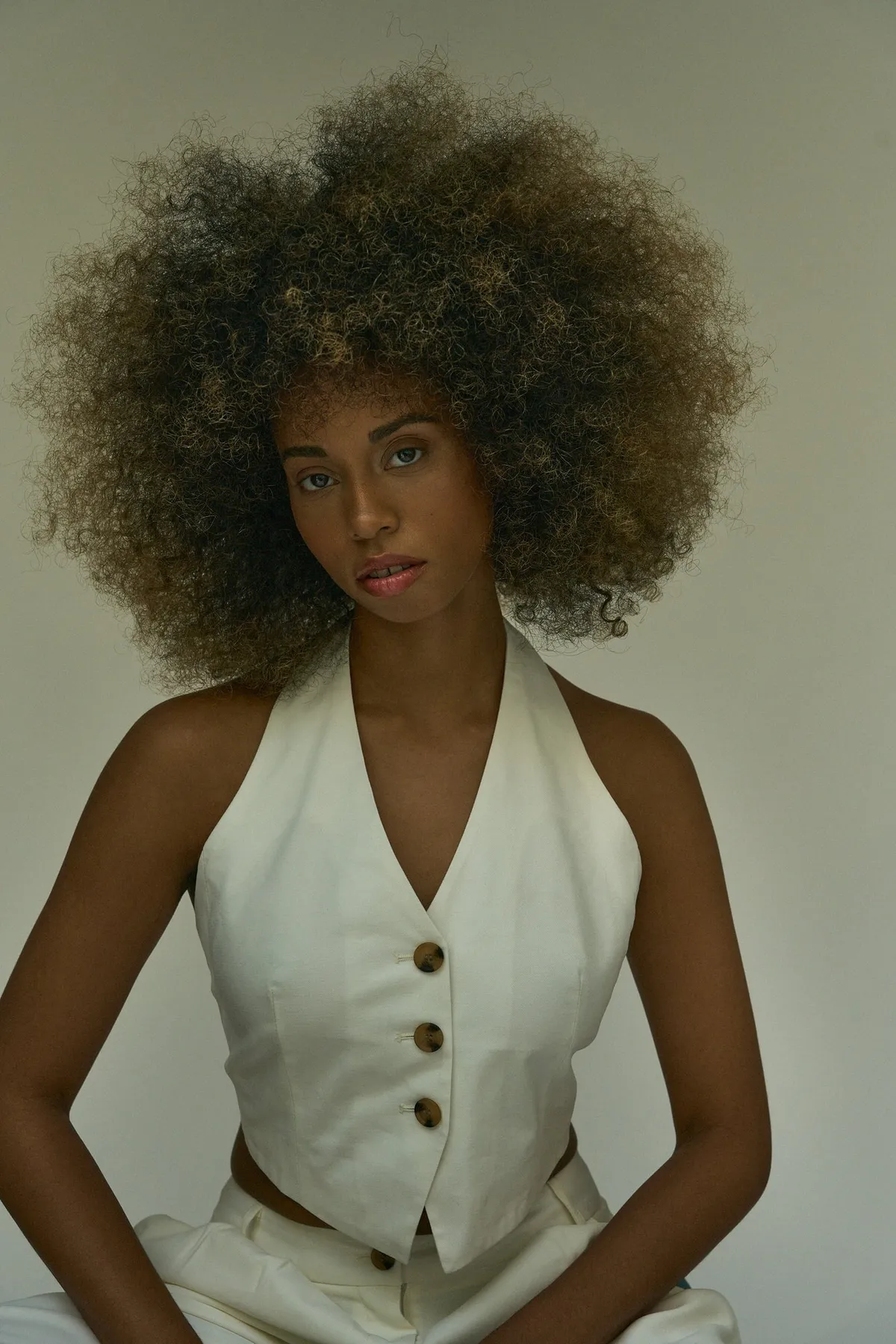 Reflections of Identity, model with a golden-brown afro in a white button waistcoat seated facing camera before olive grey, portrait series 2024, photographed by Kilian Amrehn