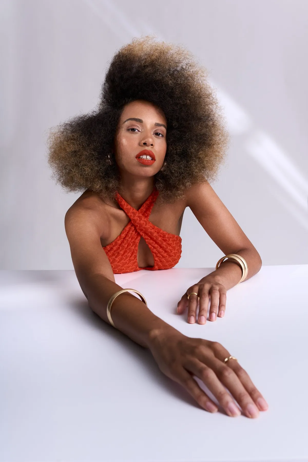 Reflections of Identity, model with red lips in an orange-red wrap top and bangles leaning on a white table, portrait series 2024, photographed by Kilian Amrehn
