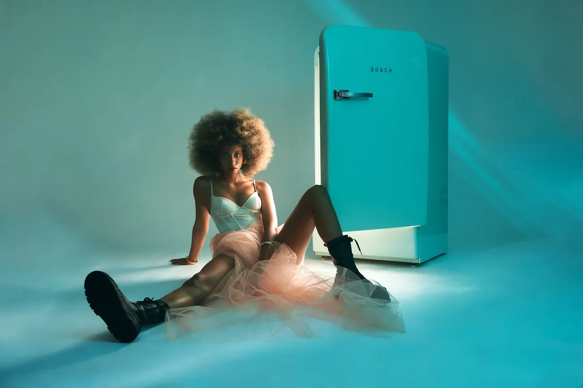 Reflections of Identity, model in a corset, tulle skirt and boots seated beside a turquoise retro fridge in teal light, portrait series 2024, photographed by Kilian Amrehn